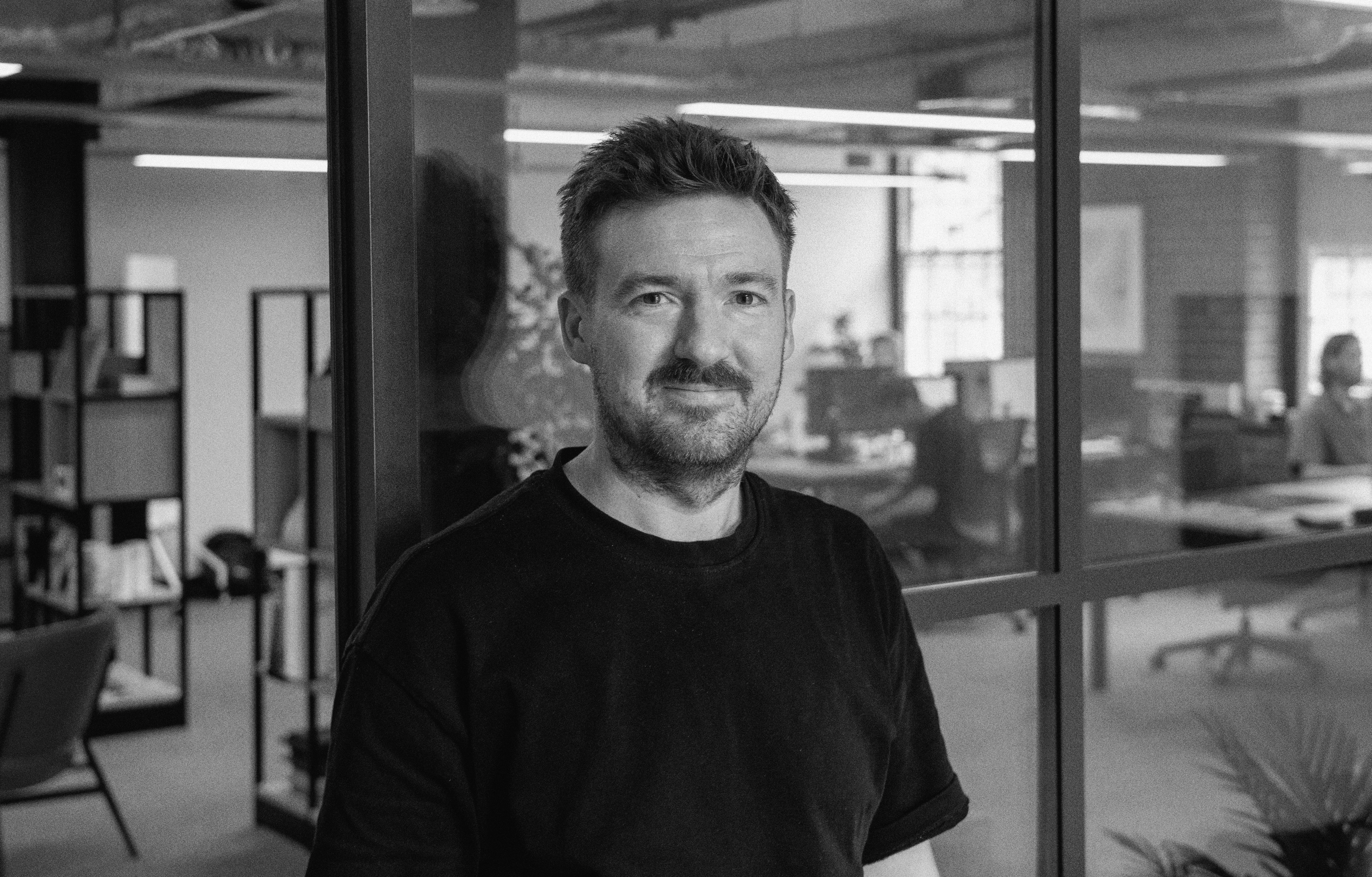 A black and white photo of James Bateson, stood against a glass partitian, with the EXP office behind him